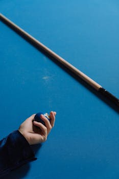 Close-up of hand holding billiard ball with cue stick on blue pool table.