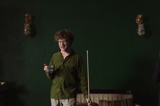 A happy young man holding whiskey and a cue stick while playing billiards indoors.