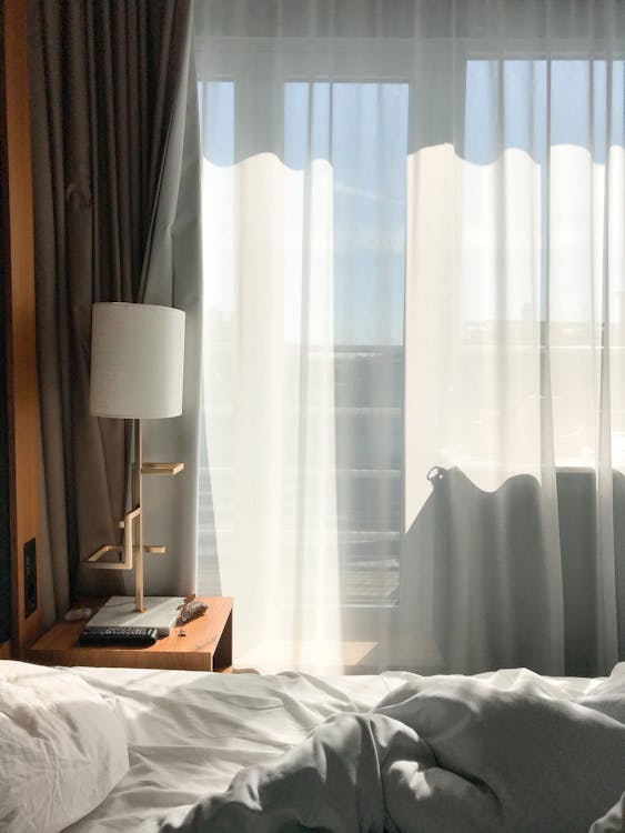 Cozy bed with crumpled blanket and pillow near nightstand with lamp and window with curtain in light apartment