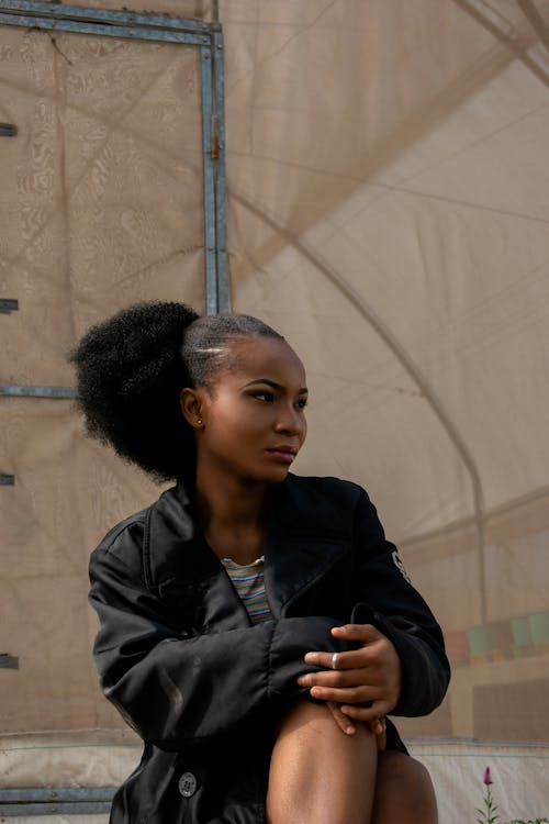 Free Confident young African American female with Afro hairstyle wearing stylish black outfit sitting on street and touching knee while looking away Stock Photo