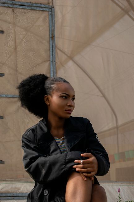 Confident young African American female with Afro hairstyle wearing stylish black outfit sitting on street and touching knee while looking away