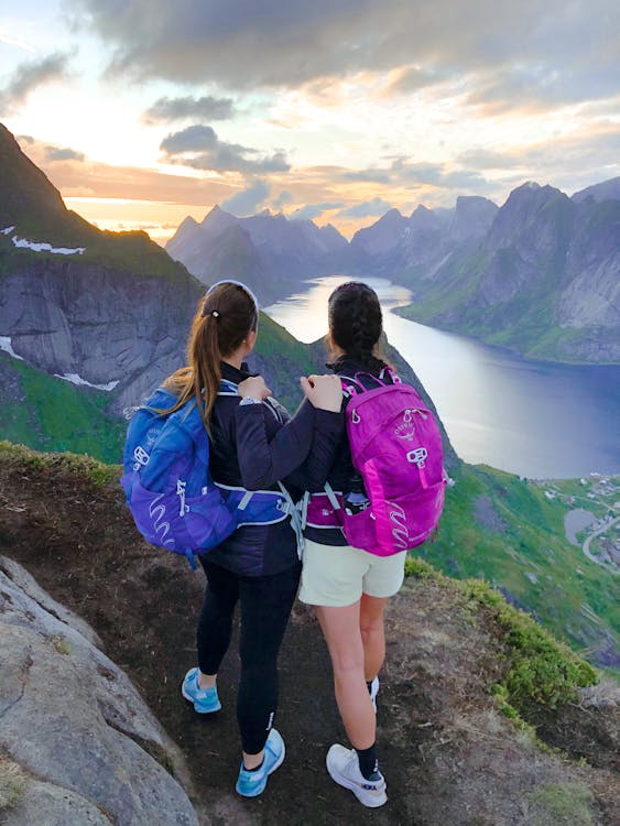 From above back view of anonymous female travelers with rucksacks embracing while contemplating mounts and pond under cloudy sky at sunset