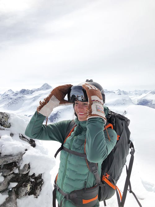 Free From above of glad female traveler with rucksack in snow goggles and outerwear standing on mount under cloudy sky while looking away in wintertime Stock Photo