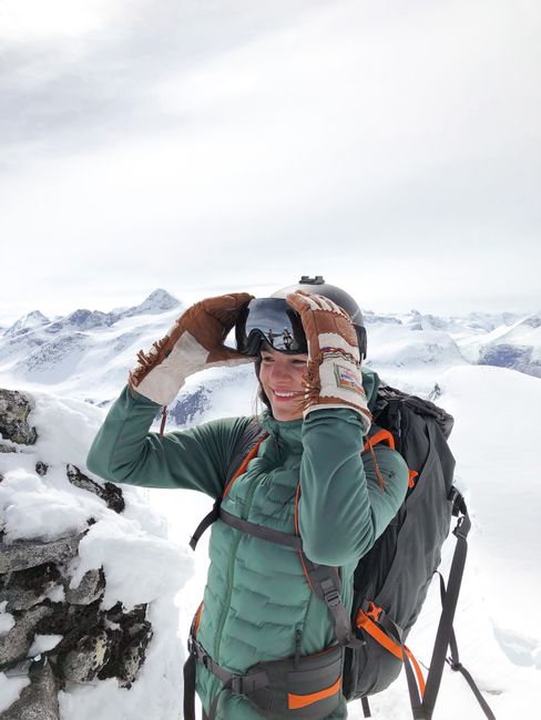 From above of glad female traveler with rucksack in snow goggles and outerwear standing on mount under cloudy sky while looking away in wintertime