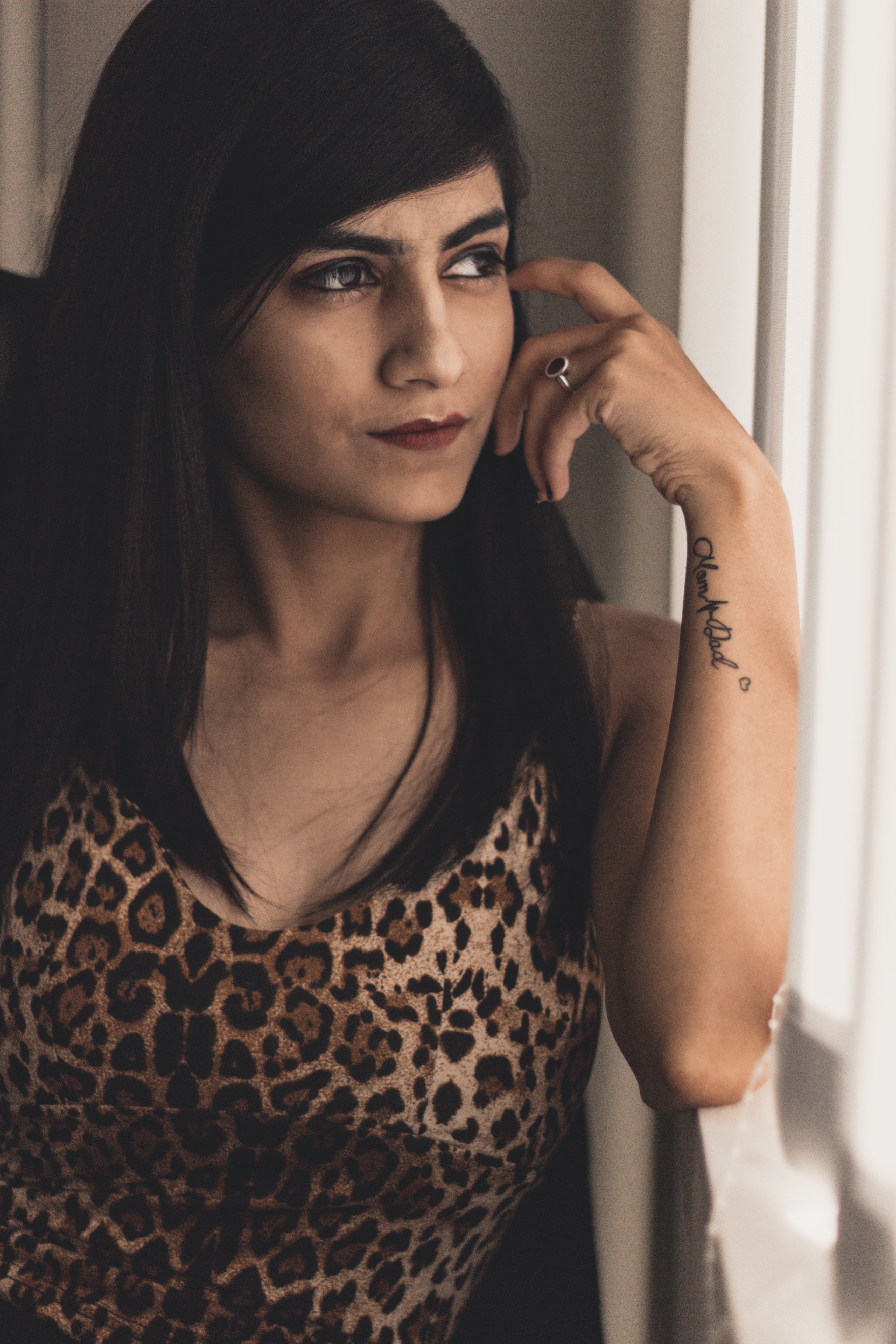 Fashionable woman in leopard print dress gazing thoughtfully out the window in soft lighting.