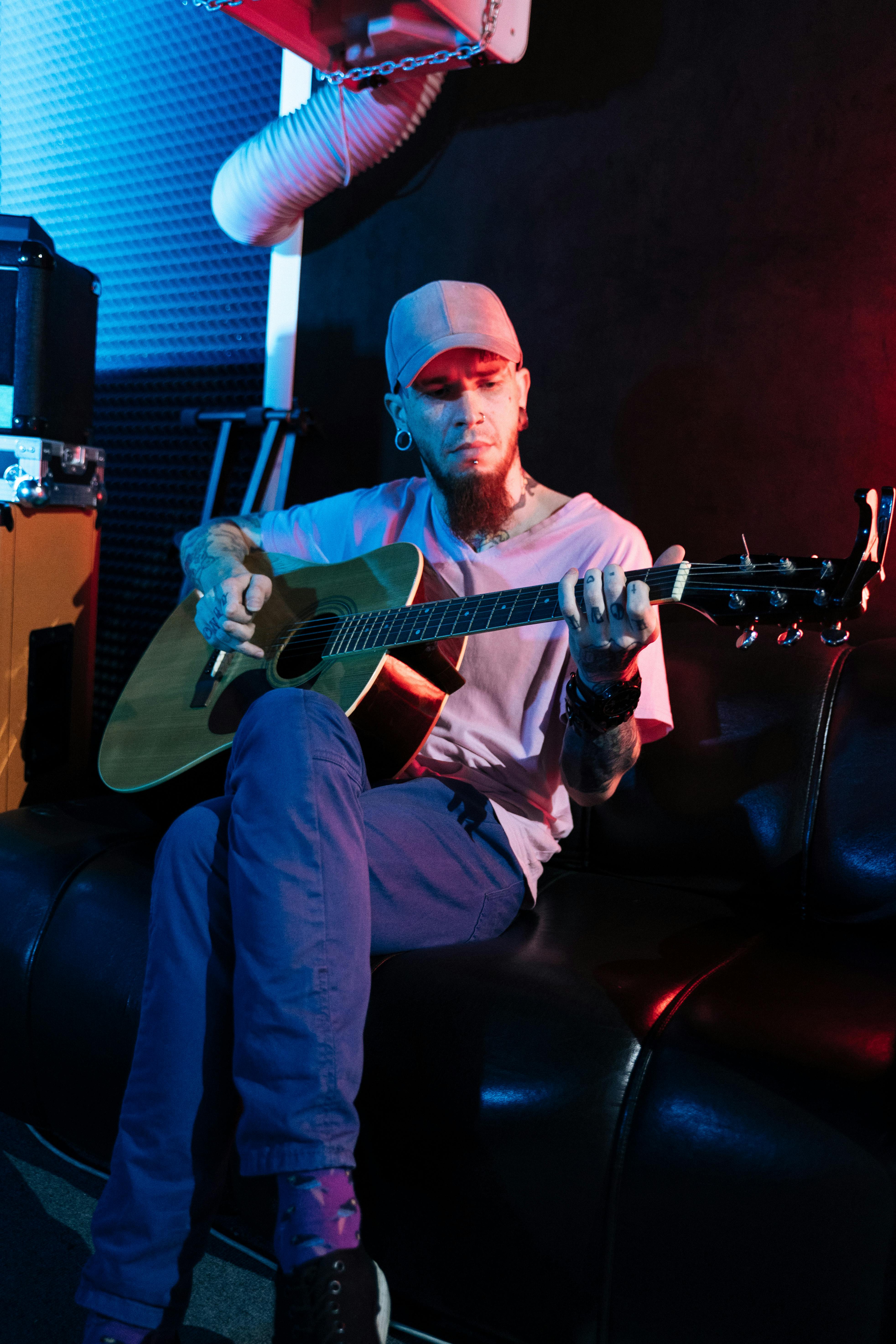 Musician with tattoos playing acoustic guitar on a sofa in a dimly lit studio.