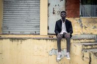 Serious young black man sitting on concrete surface near aged building with scuffed walls in daytime