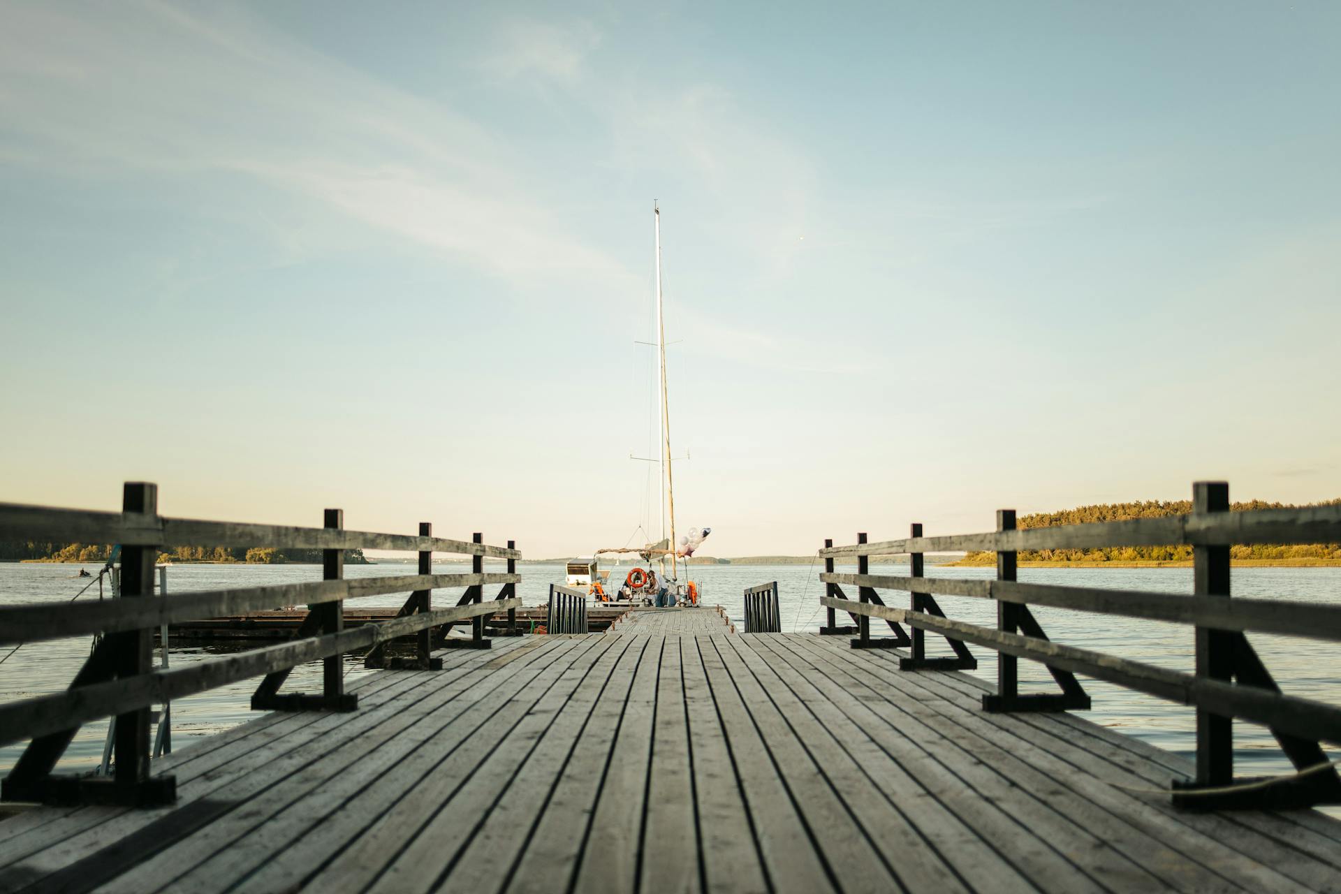 A serene scene of a wooden pier extending to a sailboat on a calm lake under a clear sky.