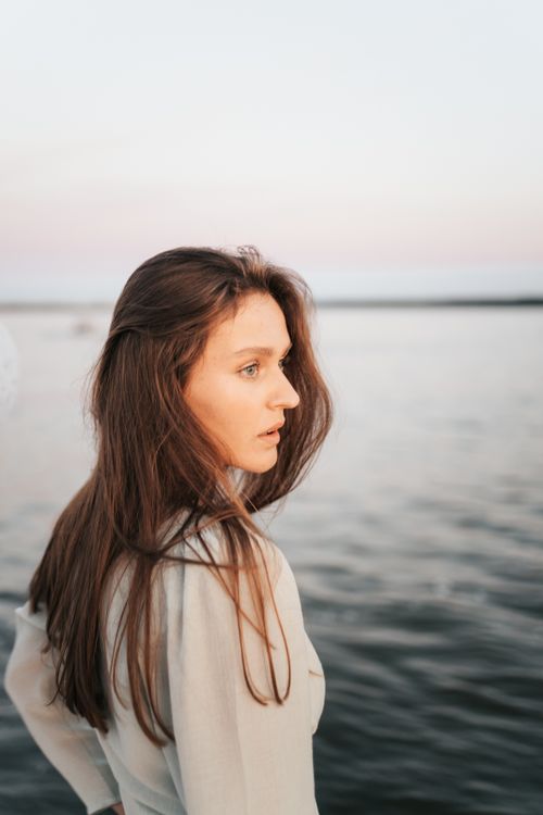 Free A young woman gazes thoughtfully at a serene lake during sunset. Stock Photo