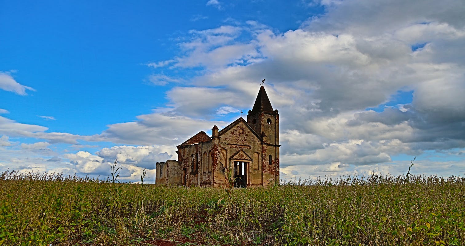 Ground level of abandoned stone church located in green grassy field against cloudy blue sky in Palmital