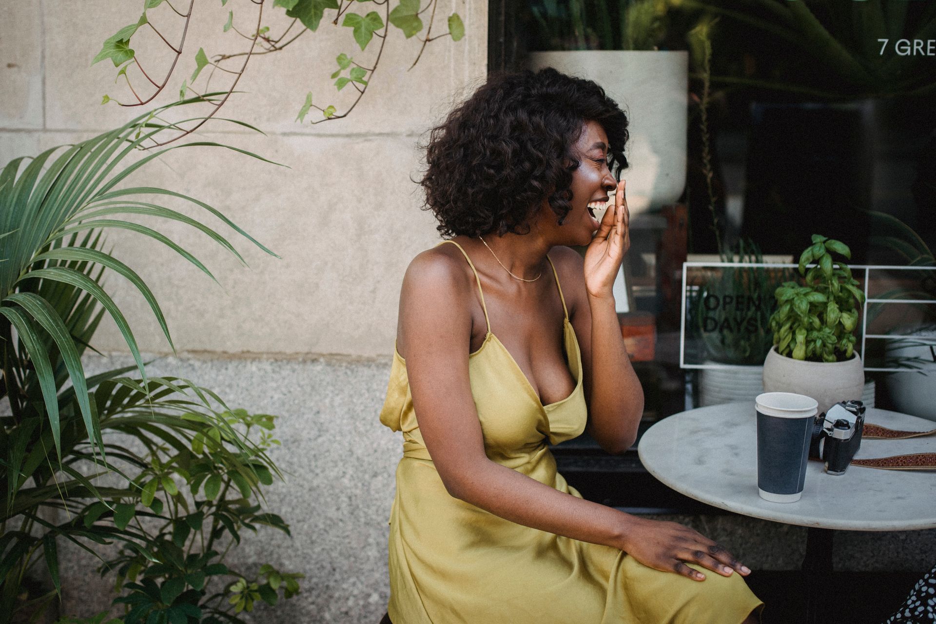 African American woman laughing in a casual outdoor cafe setting with greenery.