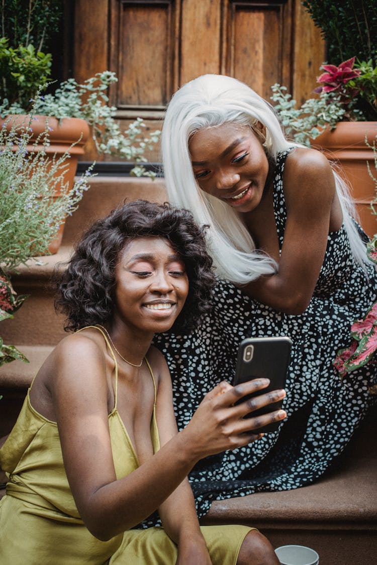 Women Sitting On Steps In Front Of A House And Looking At A Phone Screen 