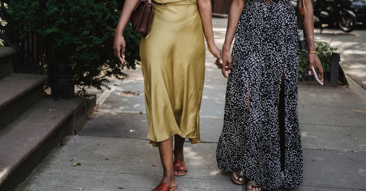 Two fashionable women in maxi dresses holding hands while walking through an urban sidewalk.