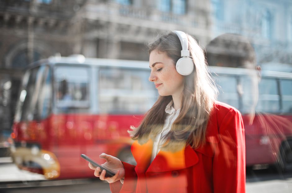Young woman in red blazer using smartphone and headphones outdoors with bus in background.