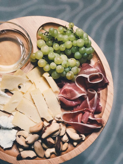From above of wooden platter served with slices of cheese and bacon near nuts and green grape with blurred background