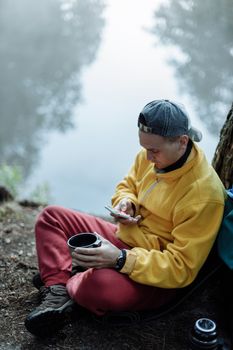 一名身穿黄色夹克的男子坐在雾湖边，用智能手机享受着户外宁静的时刻
