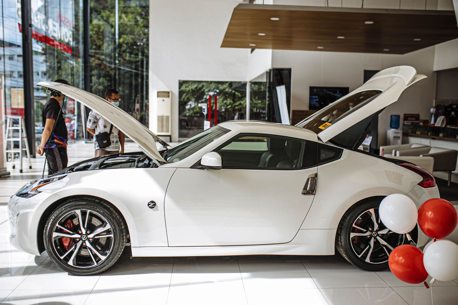 A luxurious white Nissan 370Z coupe displayed in a modern car showroom with open hoods.