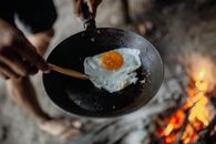 Close-up of a fried egg in a pan cooked over an open fire, showcasing rustic outdoor cooking.