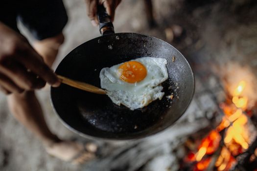 Close-up of a fried egg in a pan cooked over an open fire, showcasing rustic outdoor cooking.