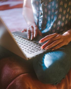 Faceless female in jeans with manicure typing on keyboard of modern laptop while sitting on bed in light room at daytime