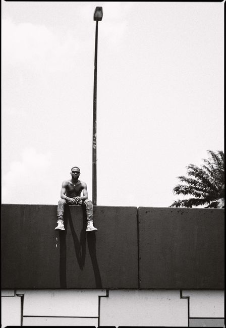 Black and white full length African American male with naked torso sitting on tall stone fence and looking at camera