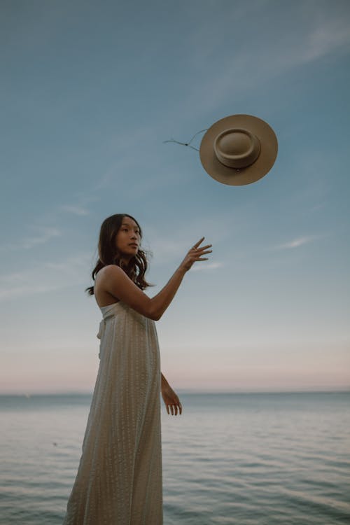 Low angle side view of romantic Asian woman in white dress throwing hat while resting near sea at sunset