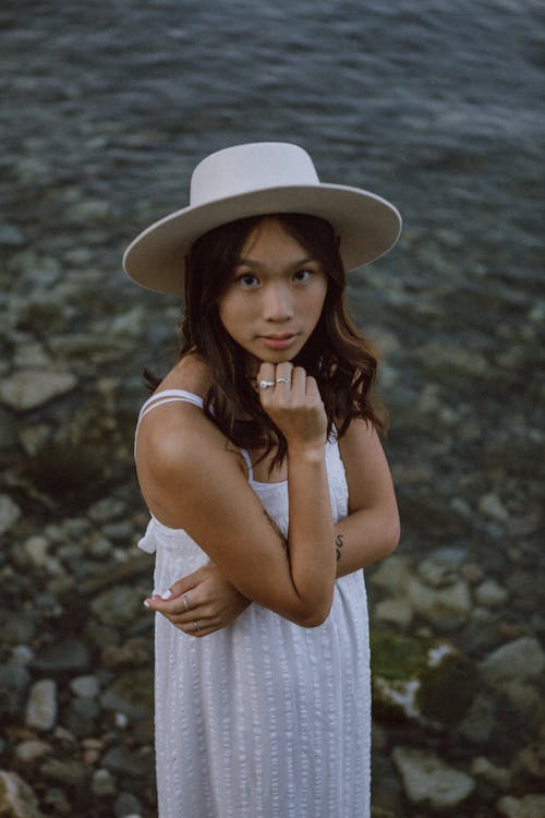 High angle of pensive young ethnic female in summer dress and hat touching chin while standing on boulders near pond