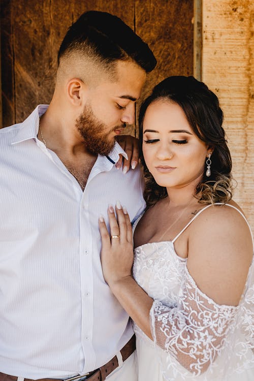 Young happy ethnic bride in white dress and groom in shirt cuddling gently during wedding celebration