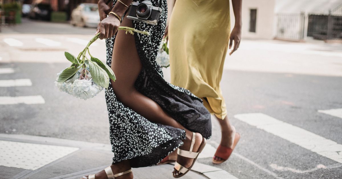 Two women in elegant dresses walk stylishly across an urban street.