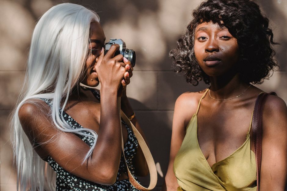 Two fashionable women captured in a sunny outdoor portrait, one using a vintage camera.