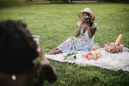 Woman in White Dress Wearing Brown Sun Hat Sitting on Green Grass While Taking Picture 