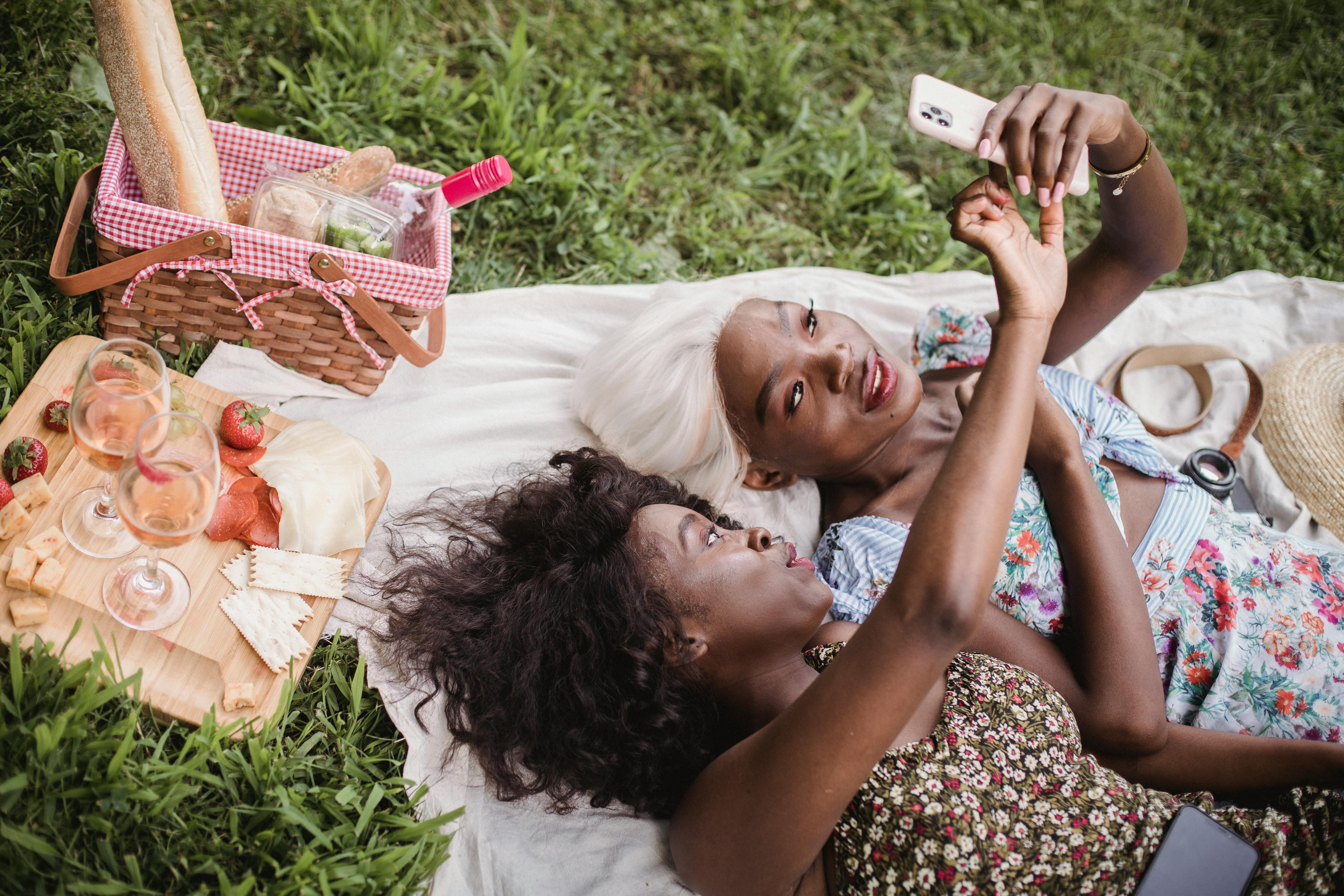 两名女子在户外野餐，自拍并品尝葡萄酒和奶酪，享受轻松时光