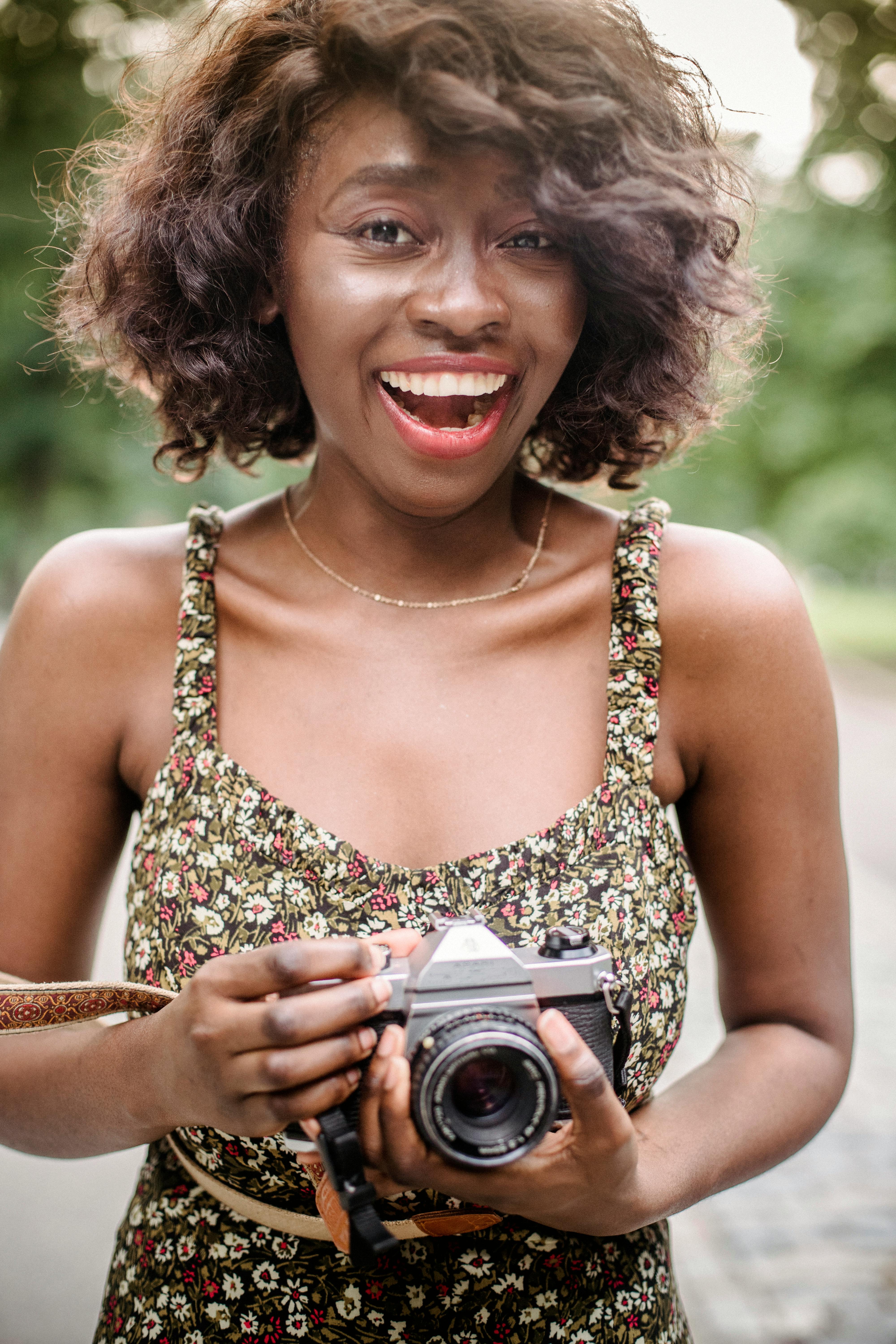 Smiling woman with curly hair and floral dress holding a camera outdoors, capturing joyful moments.