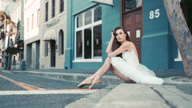 Ballet dancer in white tutu poses gracefully on a city street curb.