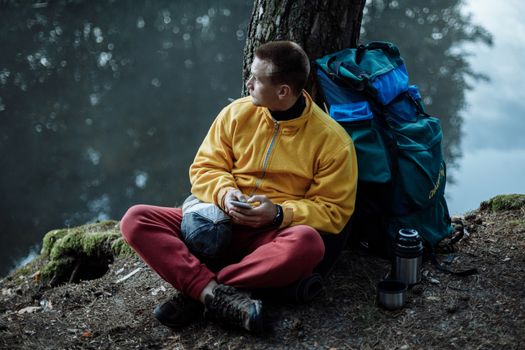 A young man in a yellow jacket rests by a tranquil lake with his backpack.