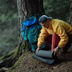 Man in a yellow fleece unrolling a dark grey sleeping mat beside a backpack under a tree