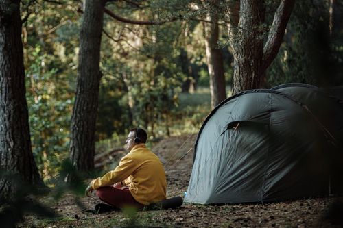 Free A person enjoying a calm morning by a tent in a forest wearing headphones. Stock Photo
