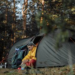 A camper in a yellow jacket sitting in the doorway of a large grey tent in a pine forest