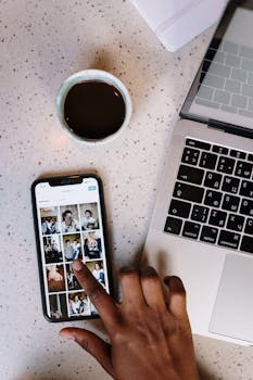 Overhead view of a person browsing photos on a smartphone beside a laptop and coffee cup in a café.