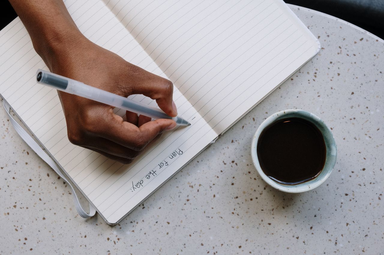 Person writing plans in a notebook beside a cup of coffee on a speckled table