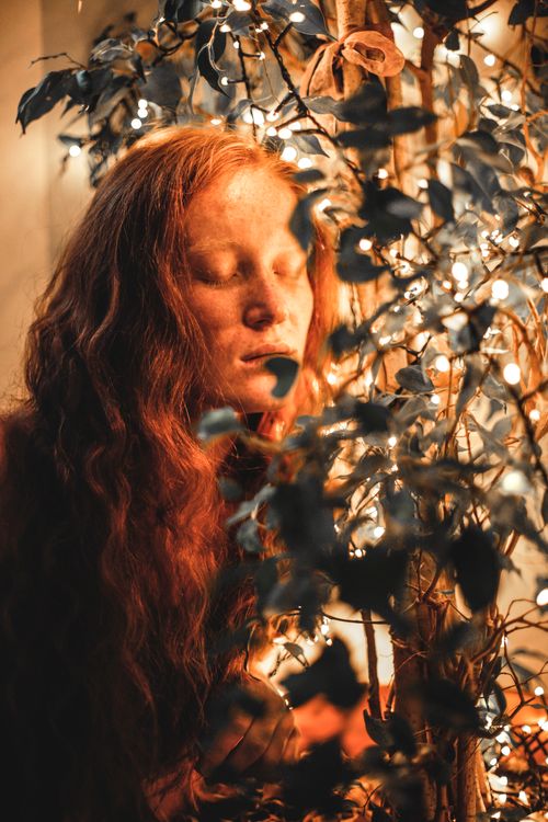 Free Dreamy female with red hair standing with closed eyes near sprigs with green foliage in room with glowing lights on background Stock Photo
