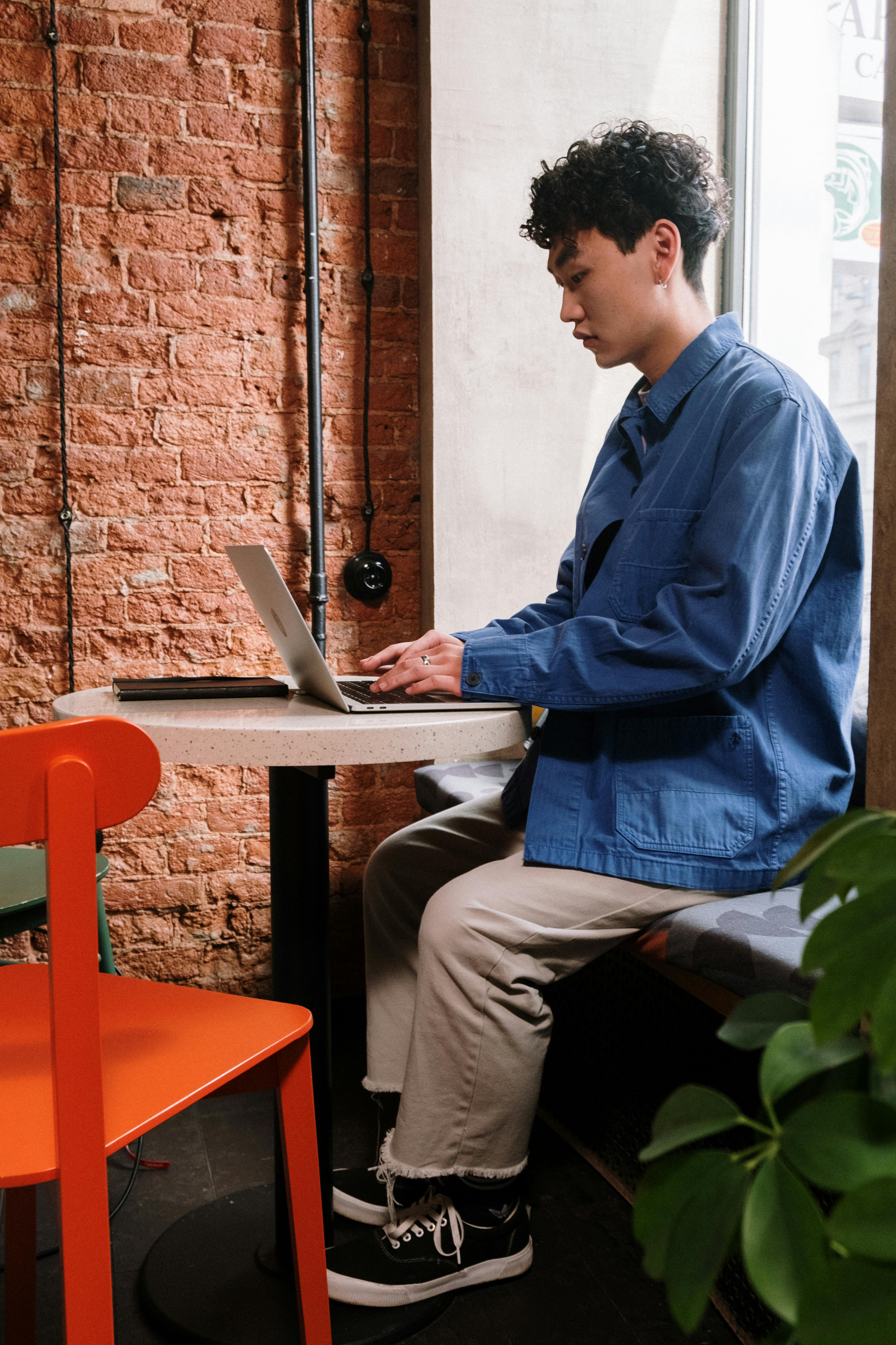 Adult male using laptop in a stylish café, brick walls, casual attire, focused on screen.