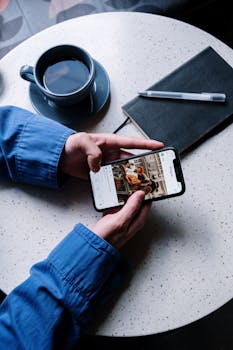 A person views Instagram on a smartphone at a cafe table with coffee, notebook, and pen.
