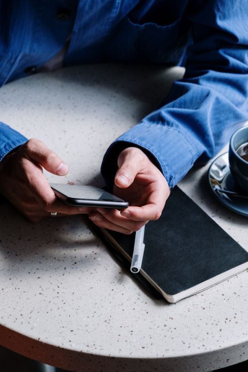 Free A person in a blue shirt using a smartphone next to a notebook and coffee on a textured table. Stock Photo