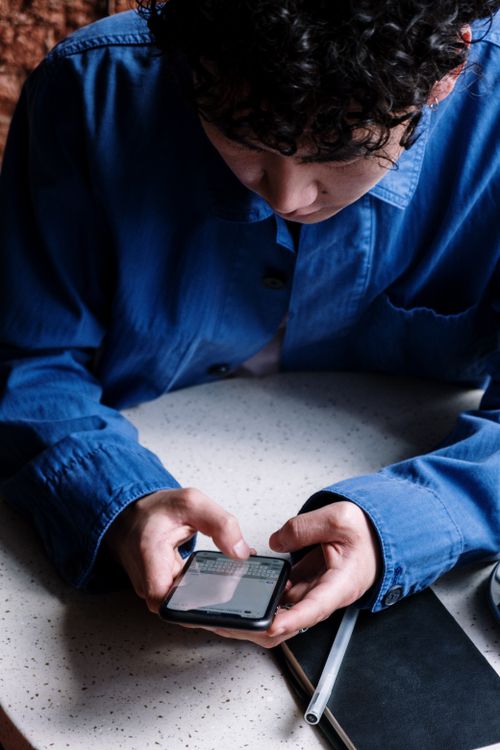 Free A young man in a blue shirt uses his smartphone in a cozy café, focusing on typing messages. Stock Photo