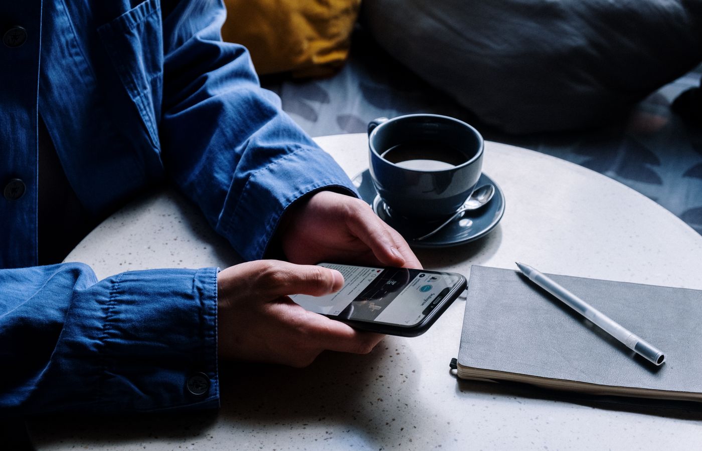 A man uses a smartphone while enjoying coffee, pen, and notepad in a cafe.