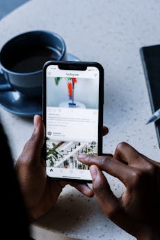 A person uses a smartphone to browse Instagram at a café table, with coffee and a notebook nearby.