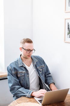 Adult man wearing glasses and a denim jacket, using a laptop indoors, focused on work.
