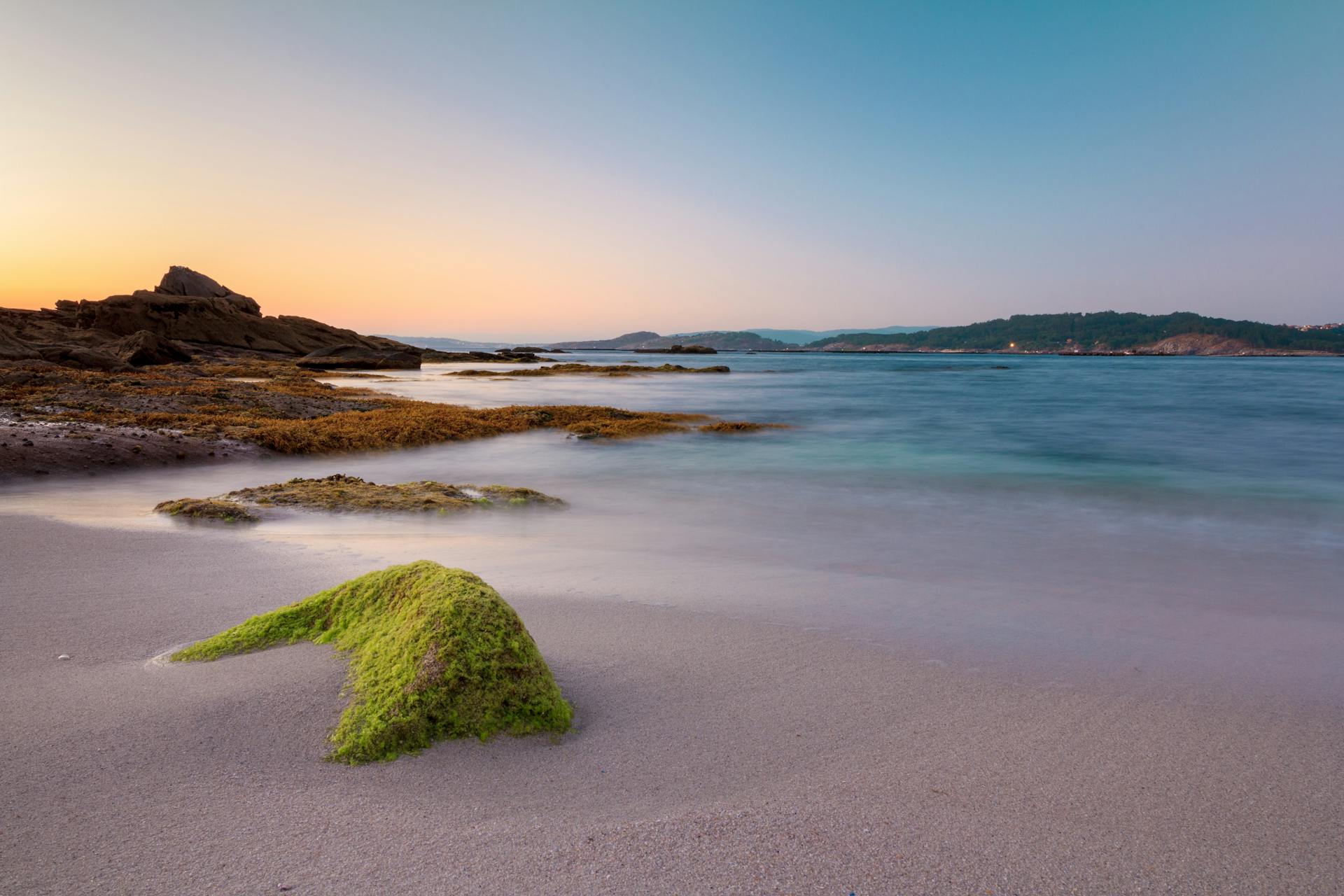 Beautiful dawn view of Aldán Beach in Spain with mossy rocks and tranquil waters.