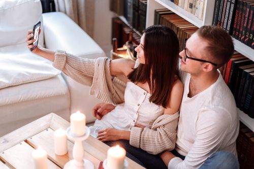 Free A happy couple taking a selfie indoors, surrounded by candles and books, creating a cozy atmosphere. Stock Photo